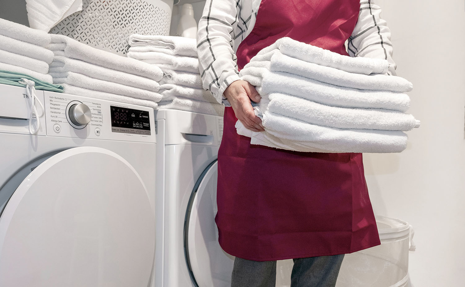 Worker in the hotel holds a lot of towels in hands near Dry machine in the laundry.